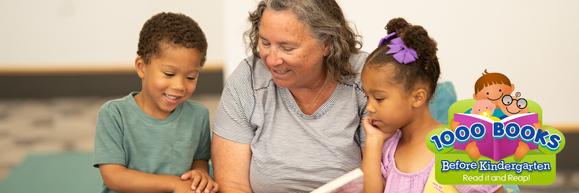 Woman and two small children reading a book in the library with the 1000 Books Before Kindergarten logo on the bottom right corner